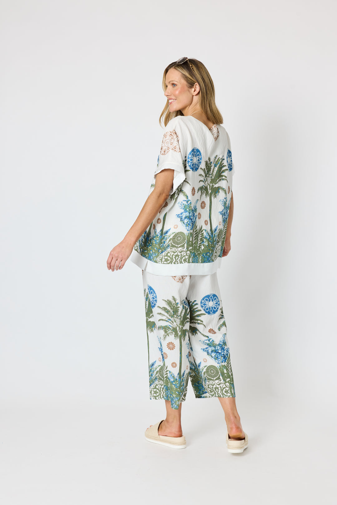 Clarity modeling a matching two-piece lounge set featuring a white short-sleeve top and wide-leg pants with a tropical palm tree and floral print in blue, green, and beige, set against a plain light gray studio background.