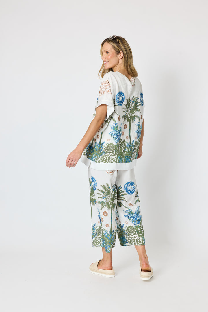 Clarity modeling a matching two-piece lounge set featuring a white short-sleeve top and wide-leg pants with a tropical palm tree and floral print in blue, green, and beige, set against a plain light gray studio background.