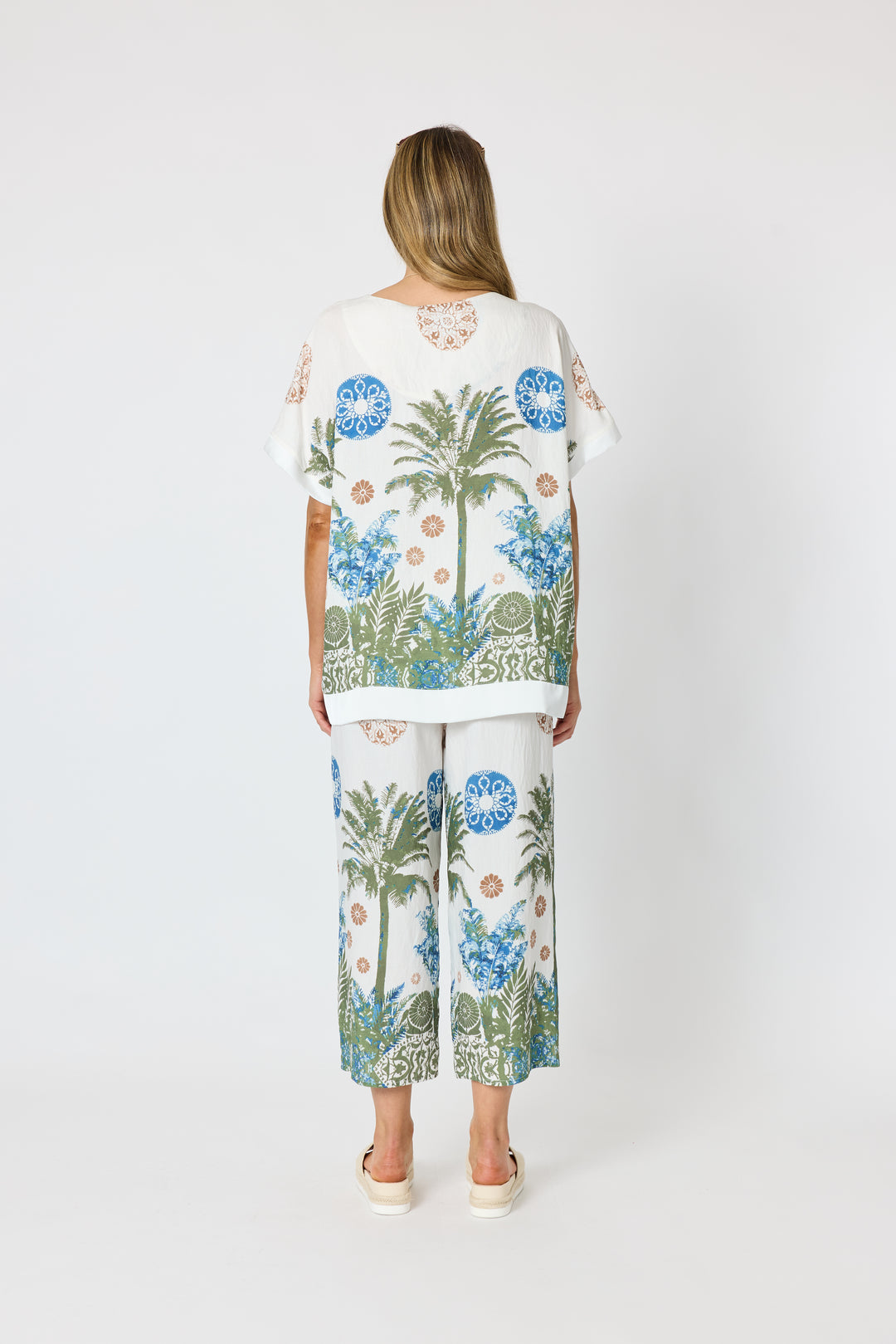 Clarity modeling a matching two-piece lounge set featuring a white base with a tropical print of palm trees, blue flowers, and green foliage, paired with white slide sandals against a light gray studio background. Back view