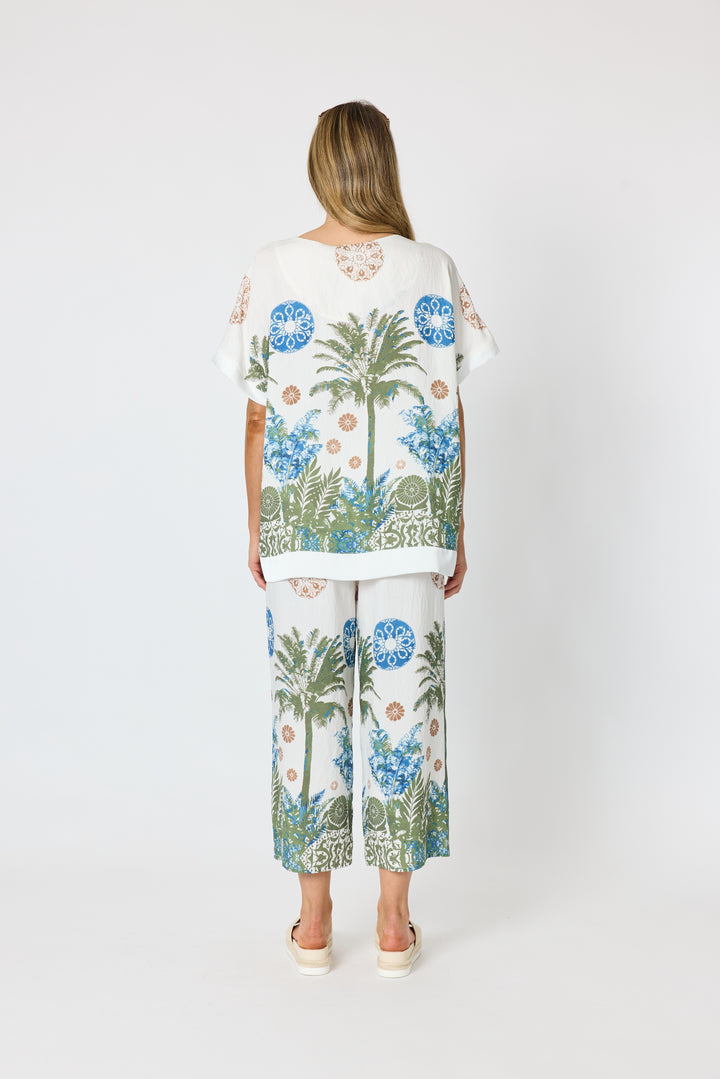 Clarity modeling a matching two-piece lounge set featuring a white short-sleeve top and wide-leg pants with a tropical palm tree and floral print in blue, green, and beige, set against a plain light gray studio background. Back view
