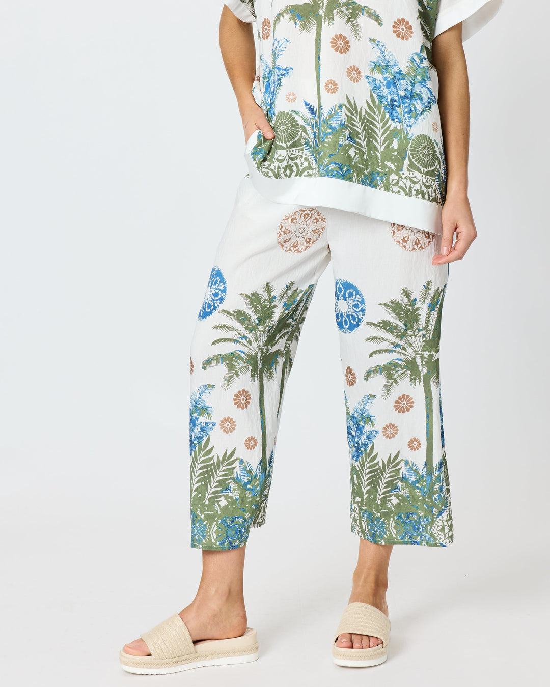 Clarity modeling a matching two-piece lounge set featuring a white base with a tropical print of palm trees, blue flowers, and green foliage, paired with white slide sandals against a light gray studio background.