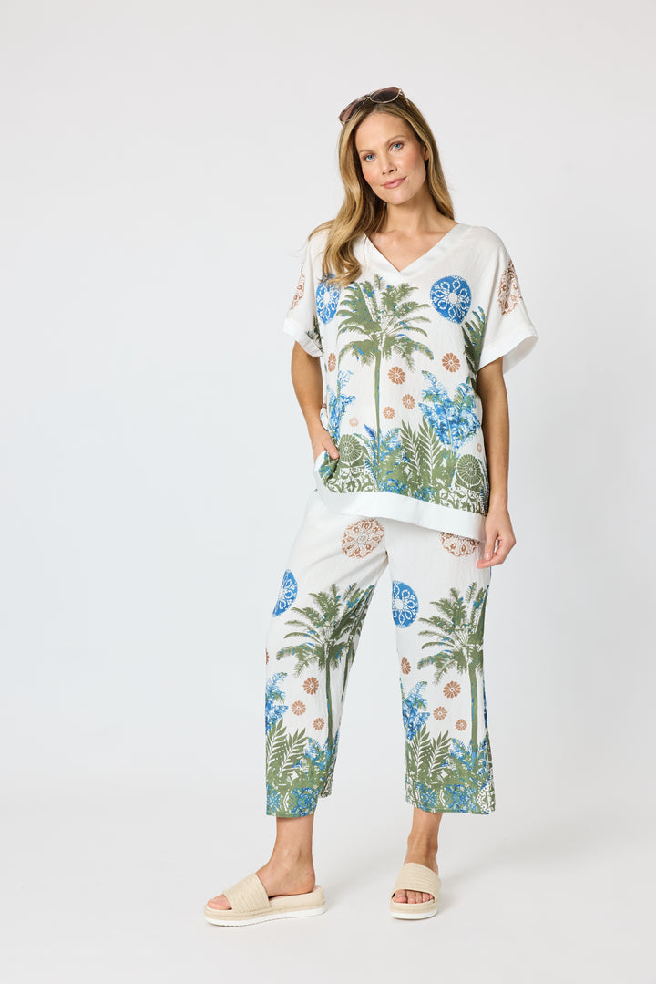 Clarity modeling a matching two-piece lounge set featuring a white short-sleeve top and wide-leg pants with a tropical palm tree and floral print in blue, green, and beige, set against a plain light gray studio background.