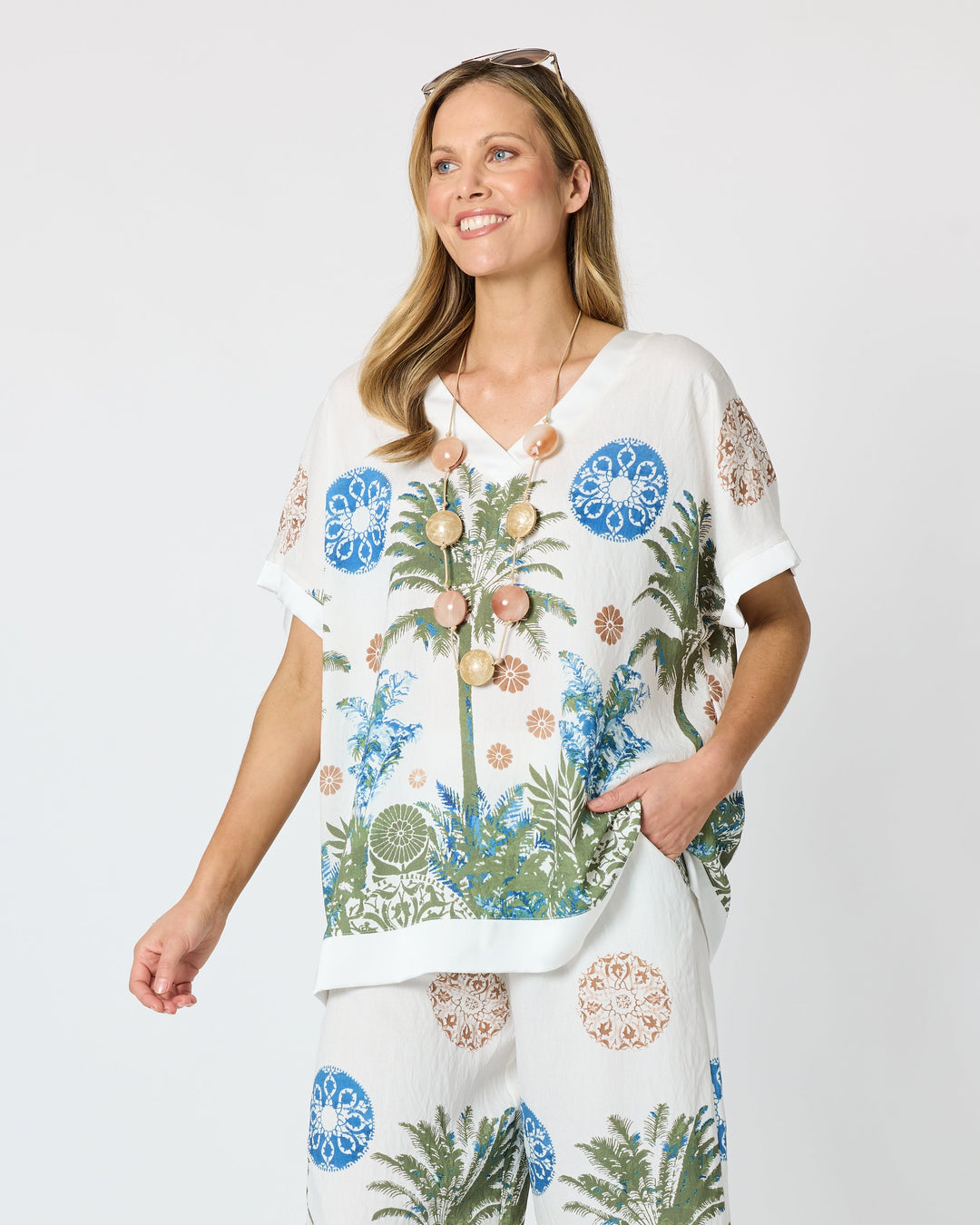 Clarity modeling a matching two-piece lounge set featuring a white short-sleeve top and wide-leg pants with a tropical palm tree and floral print in blue, green, and beige, set against a plain light gray studio background.