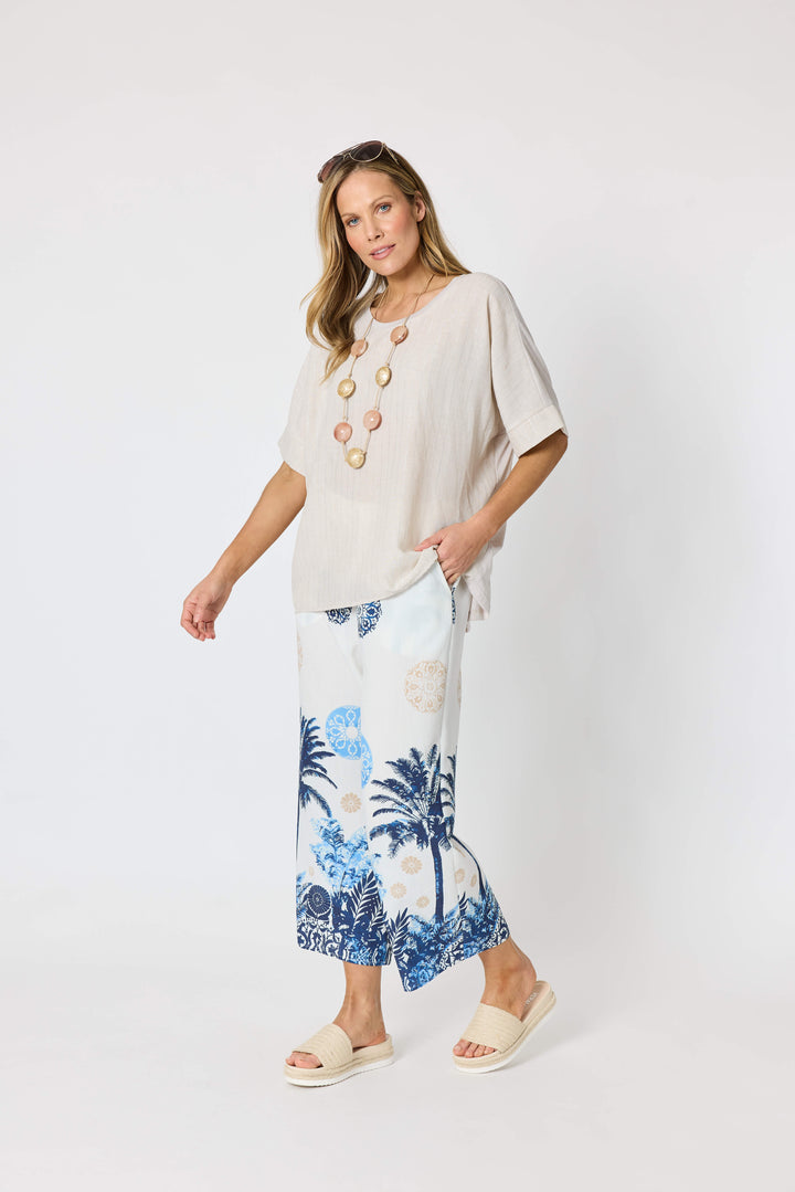 Clarity  woman modeling a two-piece lounge set consisting of a white short-sleeve top with a blue palm tree graphic and matching white wide-leg pants with a blue palm tree and beige sun motif, set against a plain white studio background.