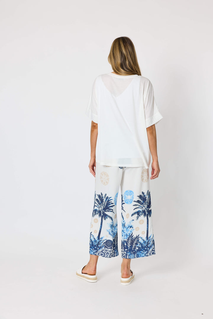 Clarity  woman modeling a two-piece lounge set consisting of a white short-sleeve top with a blue palm tree graphic and matching white wide-leg pants with a blue palm tree and beige sun motif, set against a plain white studio background.
Back view 