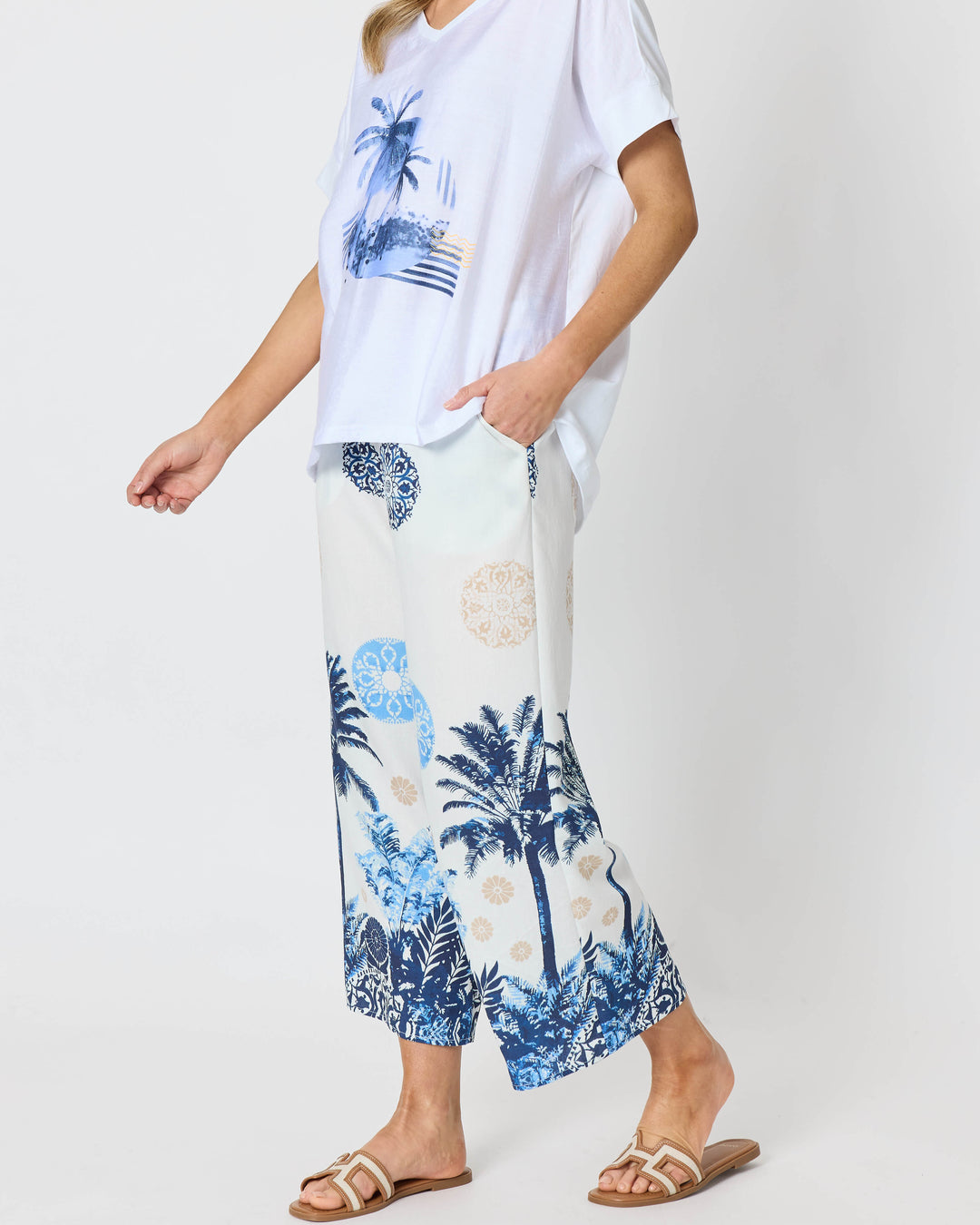 Clarity  woman modeling a two-piece lounge set consisting of a white short-sleeve top with a blue palm tree graphic and matching white wide-leg pants with a blue palm tree and beige sun motif, set against a plain white studio background.