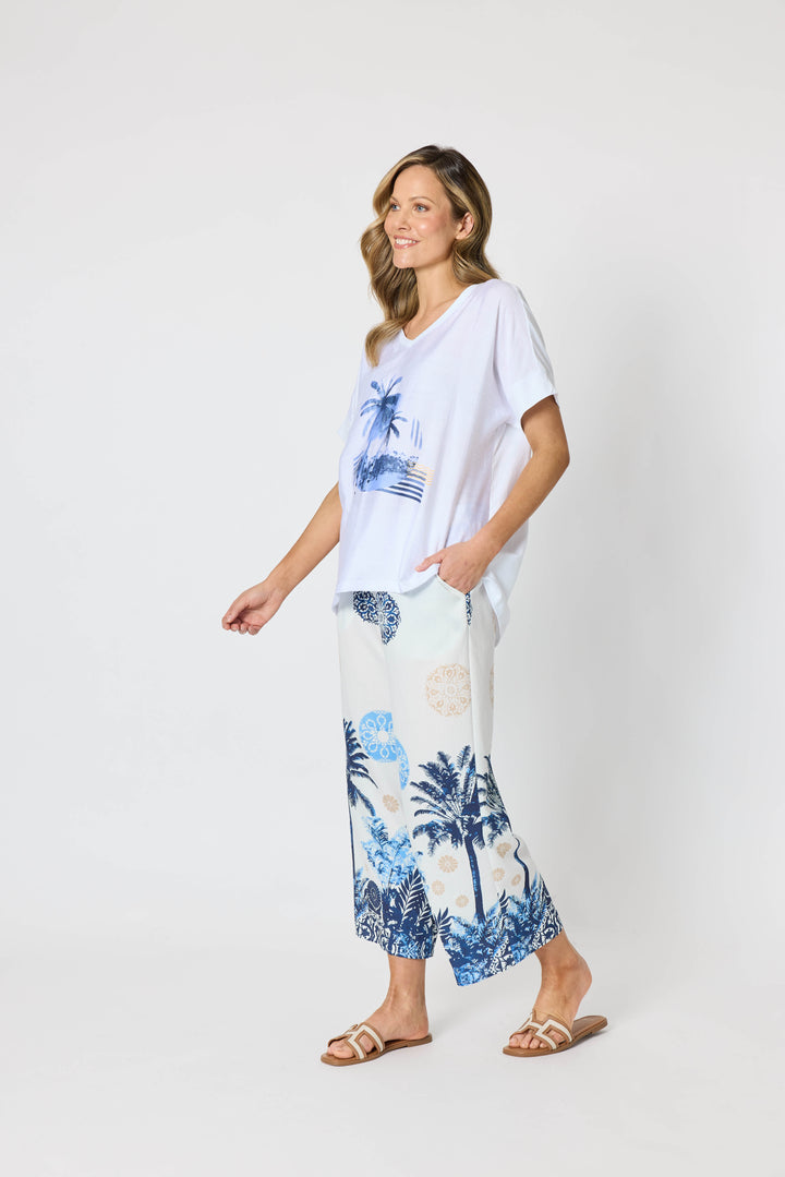 Clarity  woman modeling a two-piece lounge set consisting of a white short-sleeve top with a blue palm tree graphic and matching white wide-leg pants with a blue palm tree and beige sun motif, set against a plain white studio background.