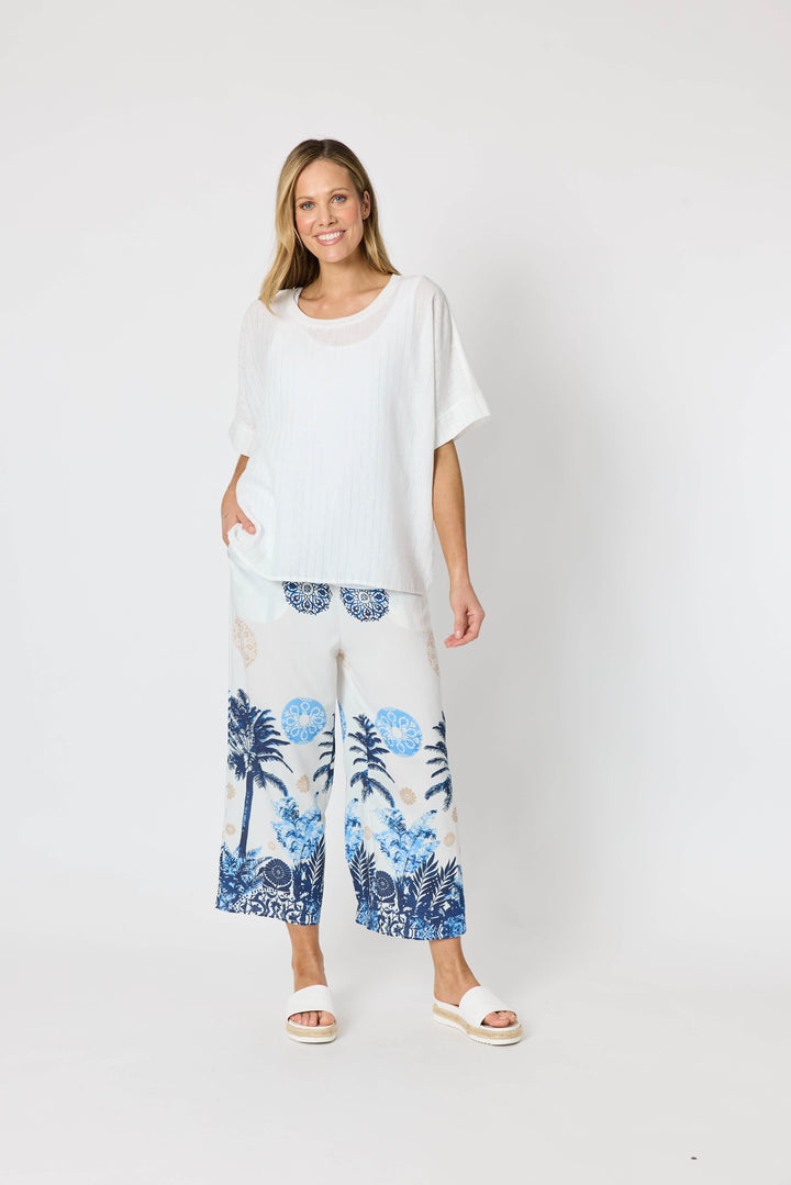 Clarity  woman modeling a two-piece lounge set consisting of a white short-sleeve top with a blue palm tree graphic and matching white wide-leg pants with a blue palm tree and beige sun motif, set against a plain white studio background.