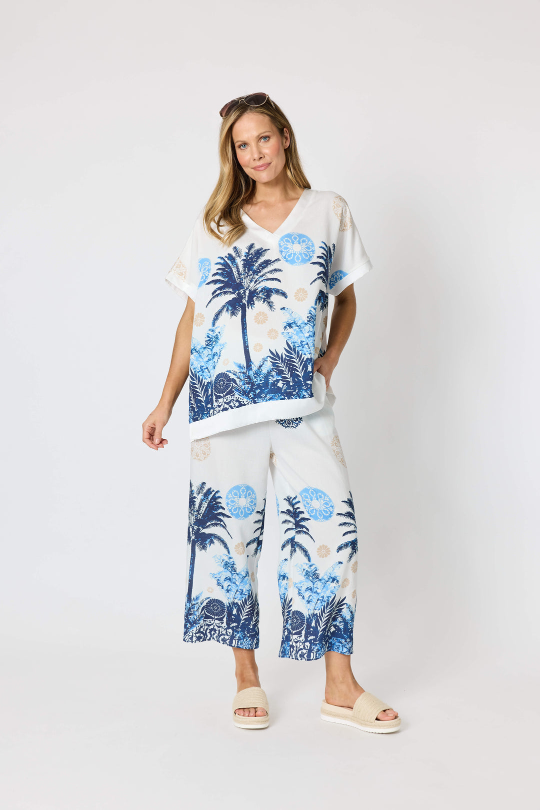 Clarity  woman modeling a two-piece lounge set consisting of a white short-sleeve top with a blue palm tree graphic and matching white wide-leg pants with a blue palm tree and beige sun motif, set against a plain white studio background.