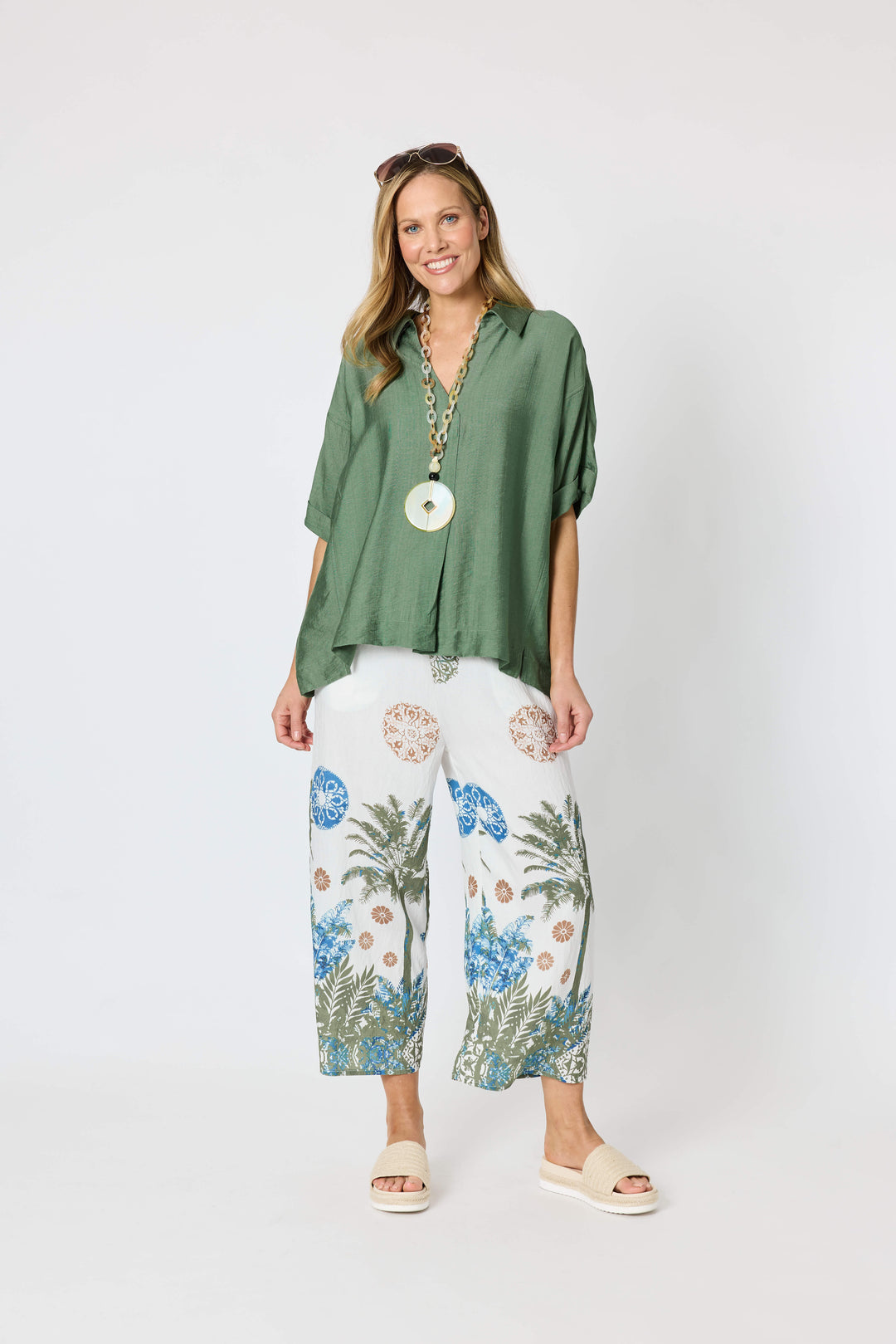 Clarity modeling a sage green short-sleeve button-up shirt with white tropical print pants, a chunky gold chain necklace with a large green disc pendant, and sunglasses on her head, set against a plain white studio background.