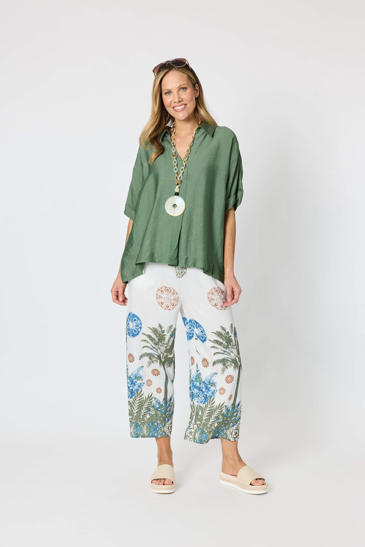 Clarity modeling a sage green short-sleeve button-up shirt with white tropical print pants, a chunky gold chain necklace with a large green disc pendant, and sunglasses on her head, set against a plain white studio background.