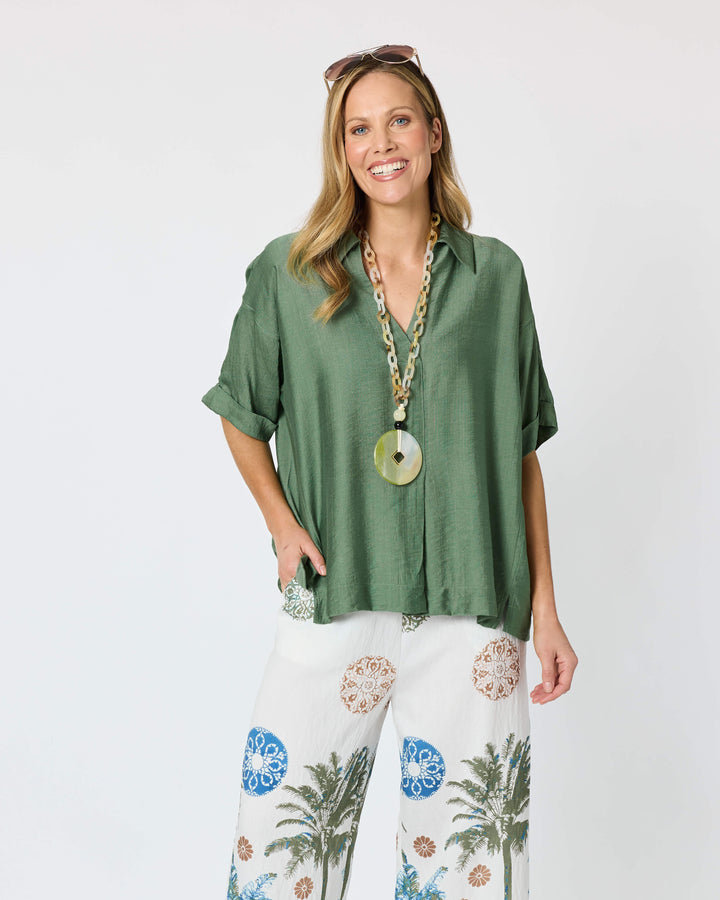 Clarity modeling a sage green short-sleeve button-up shirt with white tropical print pants, a chunky gold chain necklace with a large green disc pendant, and sunglasses on her head, set against a plain white studio background.