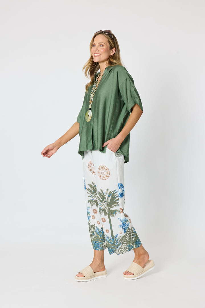Clarity modeling a sage green short-sleeve button-up shirt with white tropical print pants, a chunky gold chain necklace with a large green disc pendant, and sunglasses on her head, set against a plain white studio background.