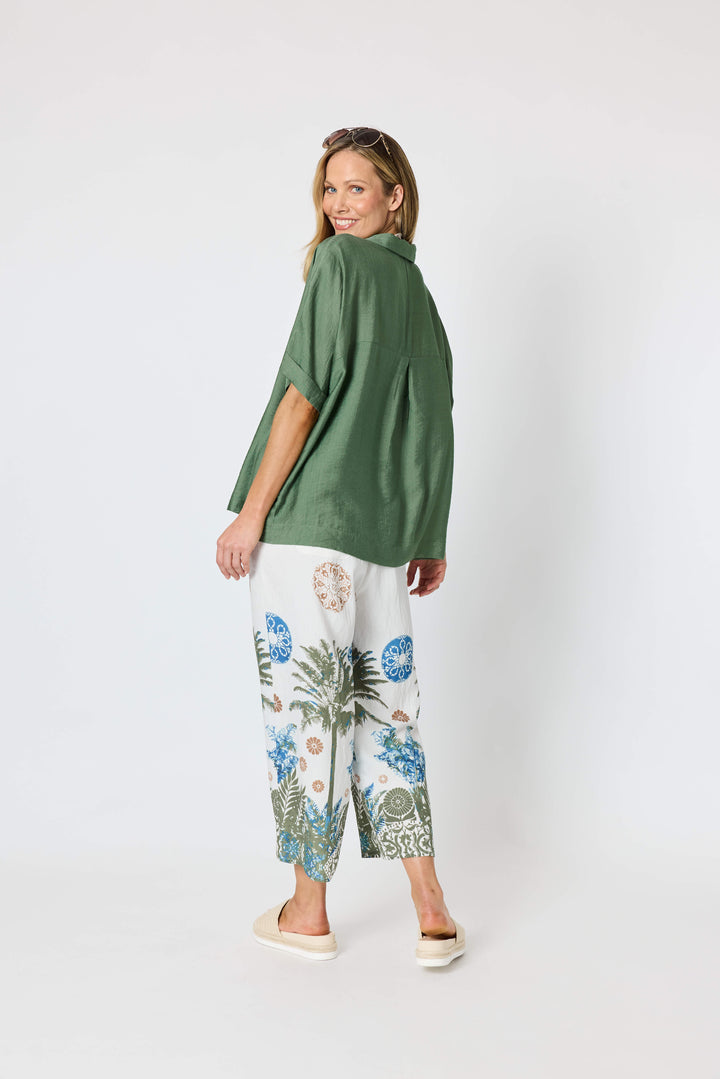 Clarity modeling a sage green short-sleeve button-up shirt with white tropical print pants, a chunky gold chain necklace with a large green disc pendant, and sunglasses on her head, set against a plain white studio background.
