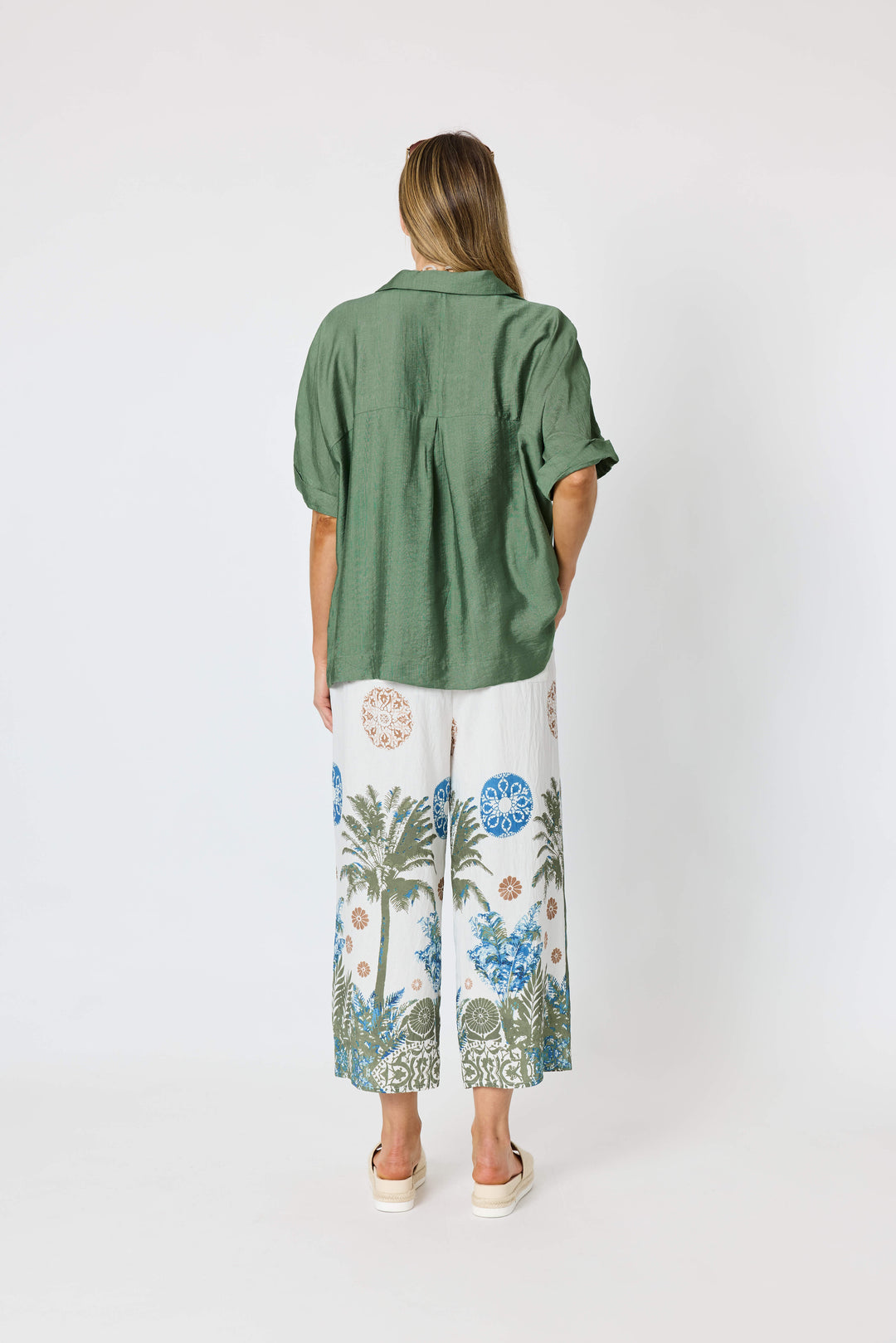 Clarity modeling a sage green short-sleeve button-up shirt with white tropical print pants, a chunky gold chain necklace with a large green disc pendant, and sunglasses on her head, set against a plain white studio background. Back view