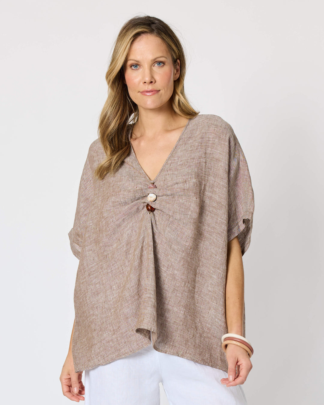 A Hammock &amp; Vine model wearing a loose taupe knit wrap top with a front knot detail, paired with white wide-leg cropped pants and tan sandals, against a plain white studio background.