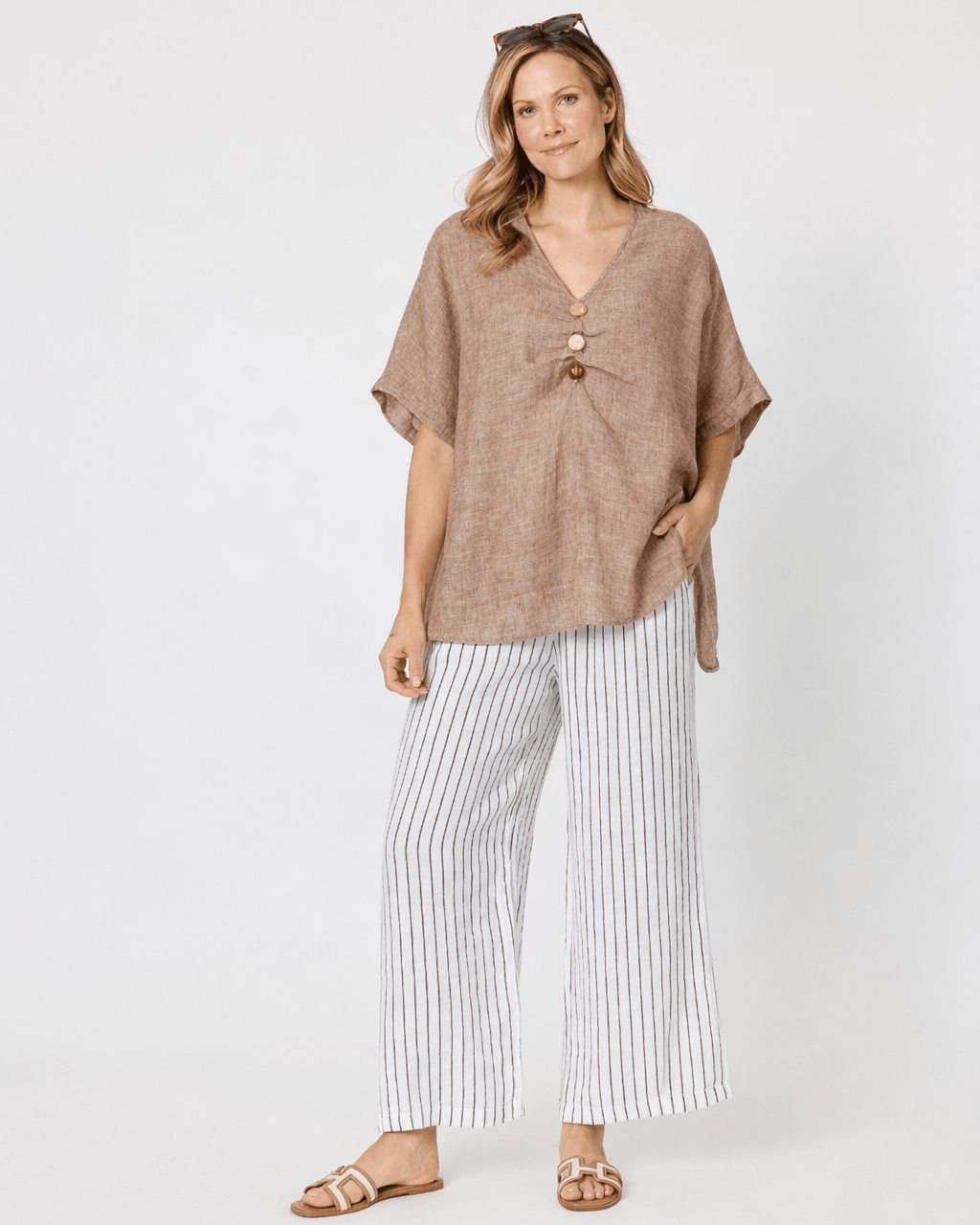 A Hammock &amp; Vine model wearing a loose taupe knit wrap top with a front knot detail, paired with white wide-leg cropped pants and tan sandals, against a plain white studio background.