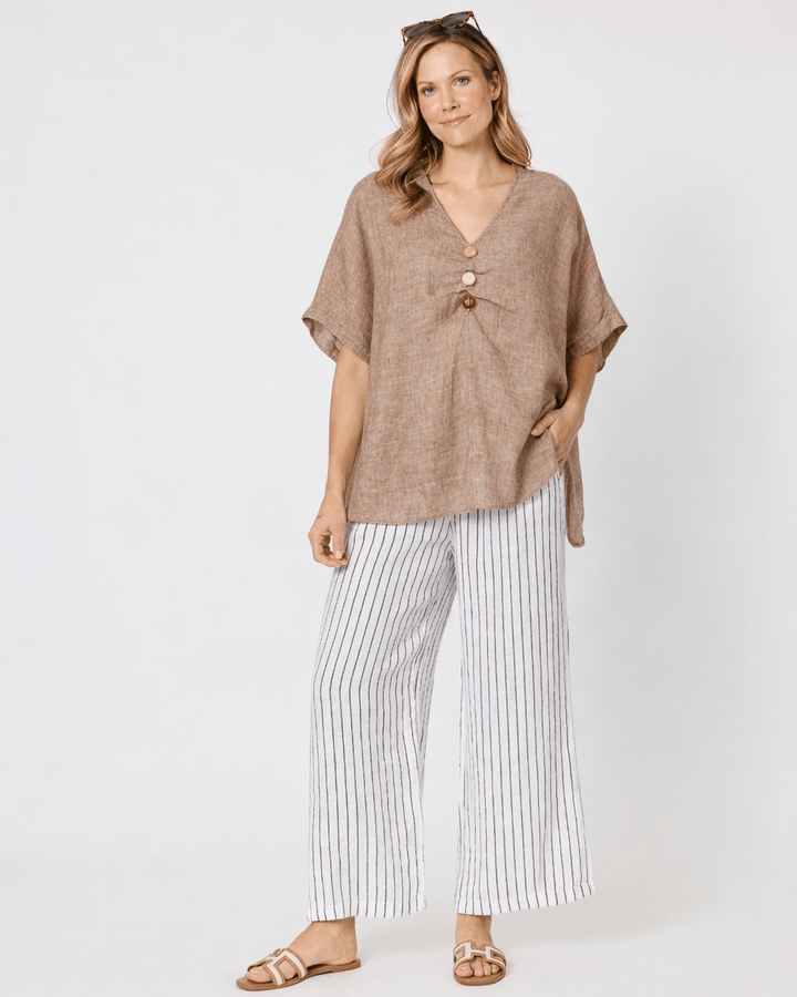 A Hammock &amp; Vine model wearing a loose taupe knit wrap top with a front knot detail, paired with white wide-leg cropped pants and tan sandals, against a plain white studio background.