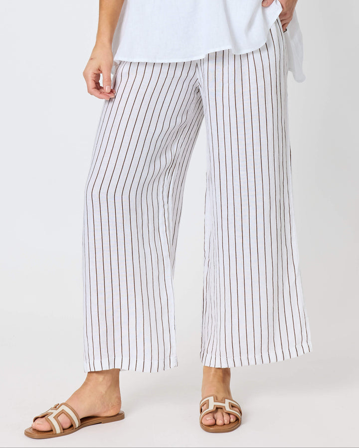 A hammock &amp; vine model wearing a white short-sleeve button-front top and white pants with thin vertical brown stripes, standing against a plain light gray studio background. 