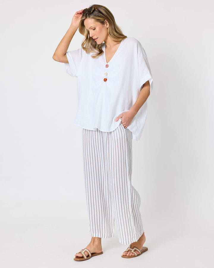 A hammock &amp; vine model wearing a white short-sleeve button-front top and white pants with thin vertical brown stripes, standing against a plain light gray studio background. 