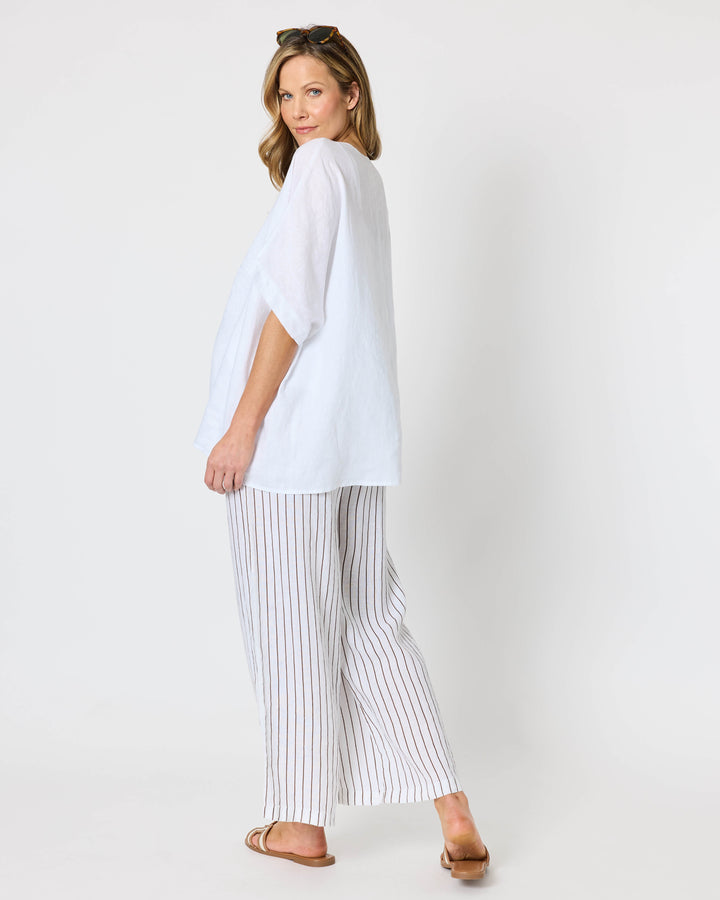 A hammock &amp; vine model wearing a white short-sleeve button-front top and white pants with thin vertical brown stripes, standing against a plain light gray studio background. 