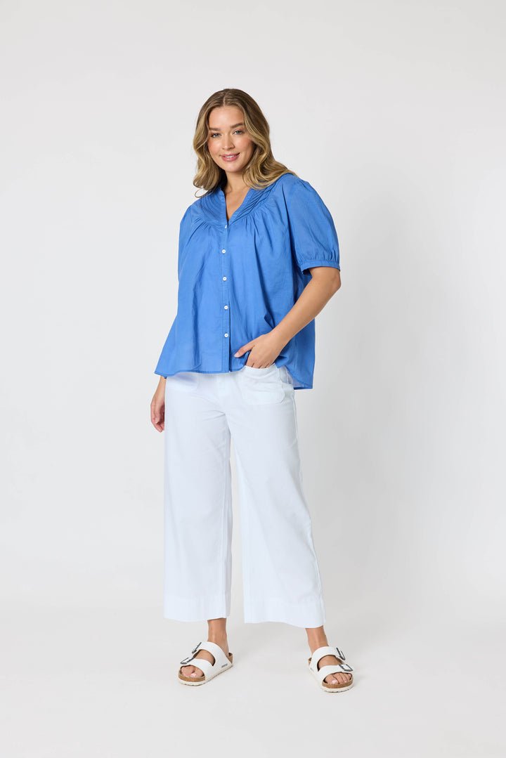 Threadz  woman modeling a blue short-sleeve button-up blouse with pleated detailing, paired with white cropped pants and white slide sandals, against a plain white studio background.