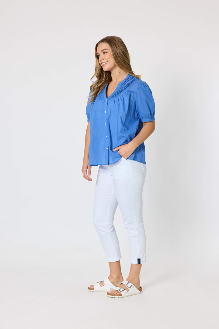 Threadz  woman modeling a blue short-sleeve button-up blouse with pleated detailing, paired with white cropped pants and white slide sandals, against a plain white studio background.