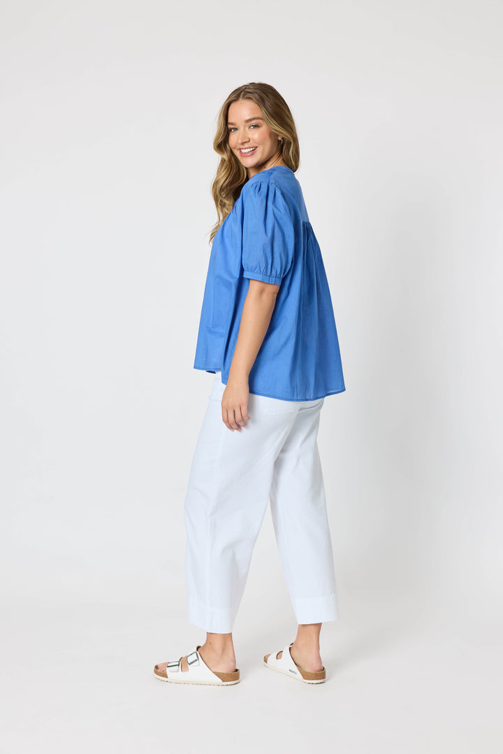 Threadz  woman modeling a blue short-sleeve button-up blouse with pleated detailing, paired with white cropped pants and white slide sandals, against a plain white studio background. Side view 