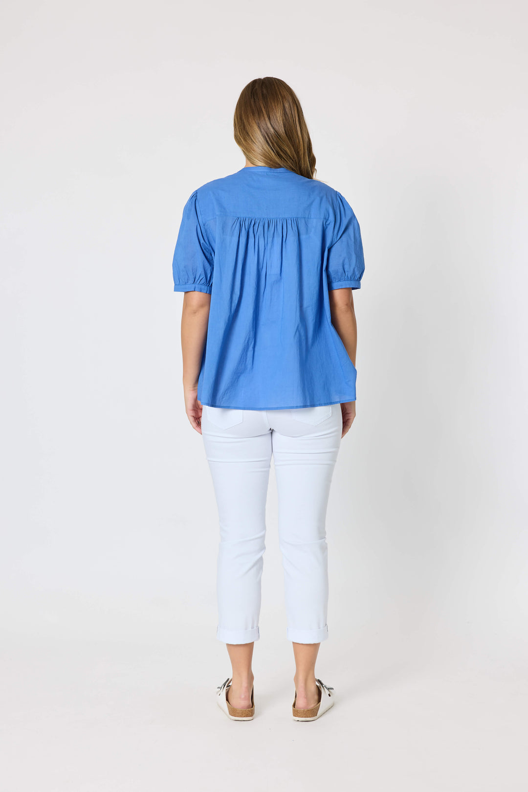 Threadz  woman modeling a blue short-sleeve button-up blouse with pleated detailing, paired with white cropped pants and white slide sandals, against a plain white studio background. Back view