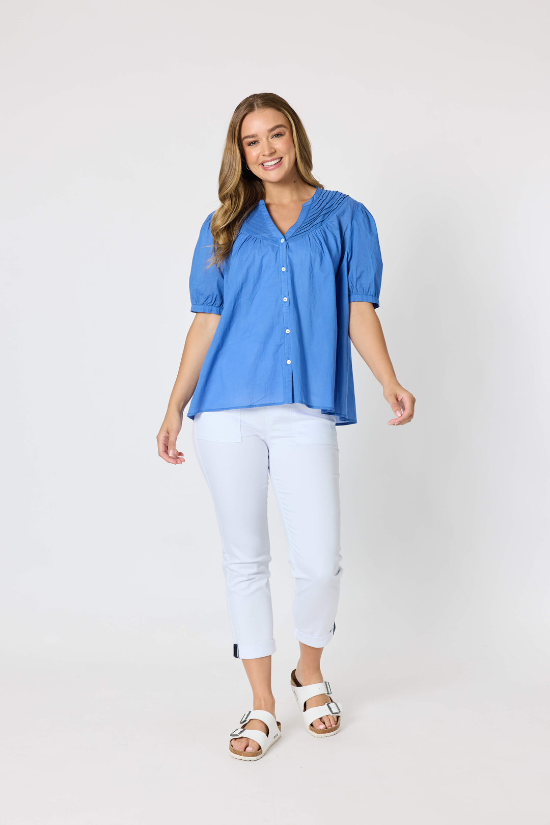 Threadz  woman modeling a blue short-sleeve button-up blouse with pleated detailing, paired with white cropped pants and white slide sandals, against a plain white studio background.