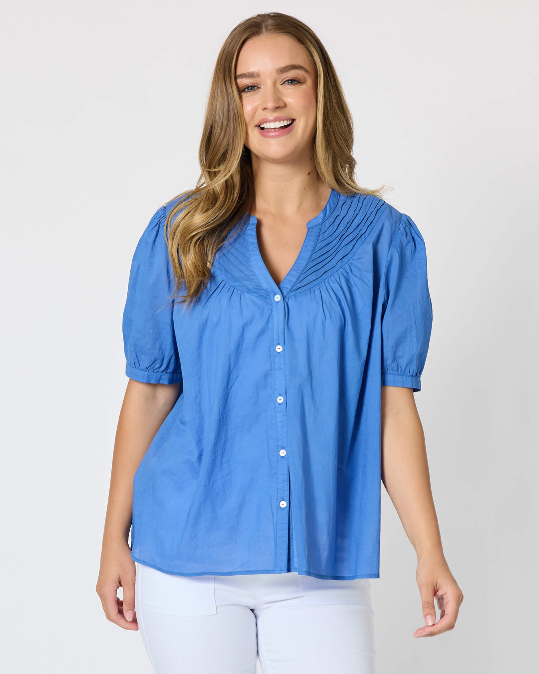 Threadz  woman modeling a blue short-sleeve button-up blouse with pleated detailing, paired with white cropped pants and white slide sandals, against a plain white studio background.