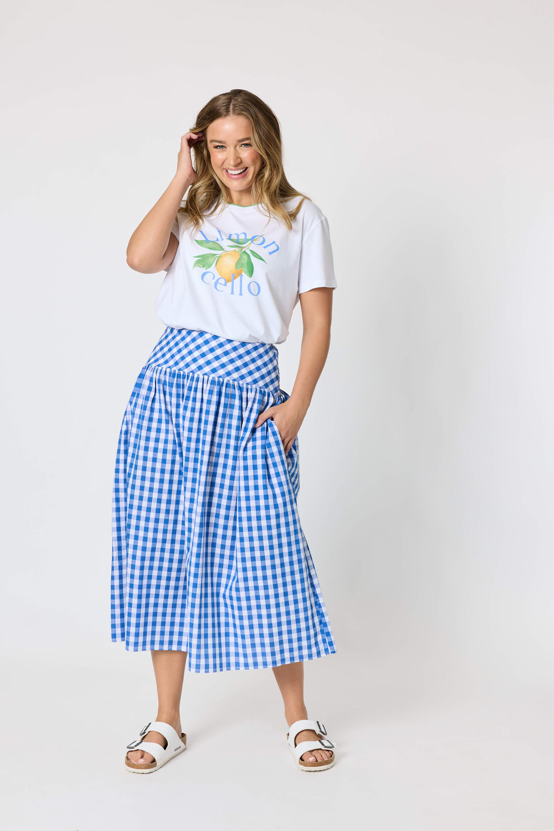 Threadz woman modeling a white graphic t-shirt and a blue and white gingham pleated midi skirt, paired with white slide sandals, against a plain white studio background.
