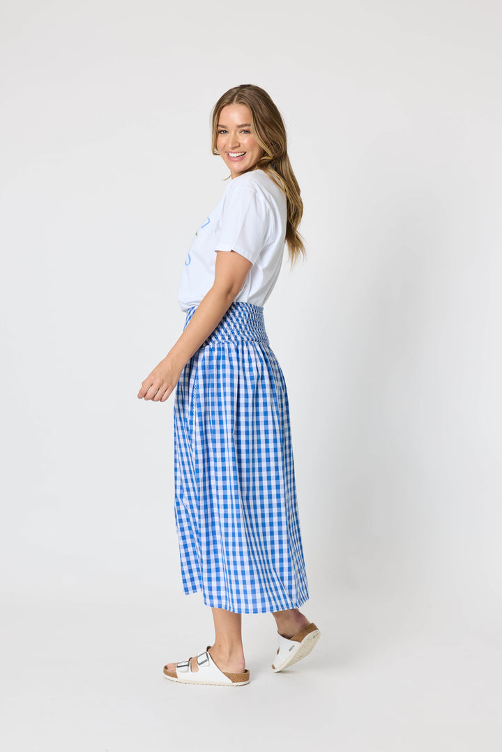 Threadz woman modeling a white graphic t-shirt and a blue and white gingham pleated midi skirt, paired with white slide sandals, against a plain white studio background.
