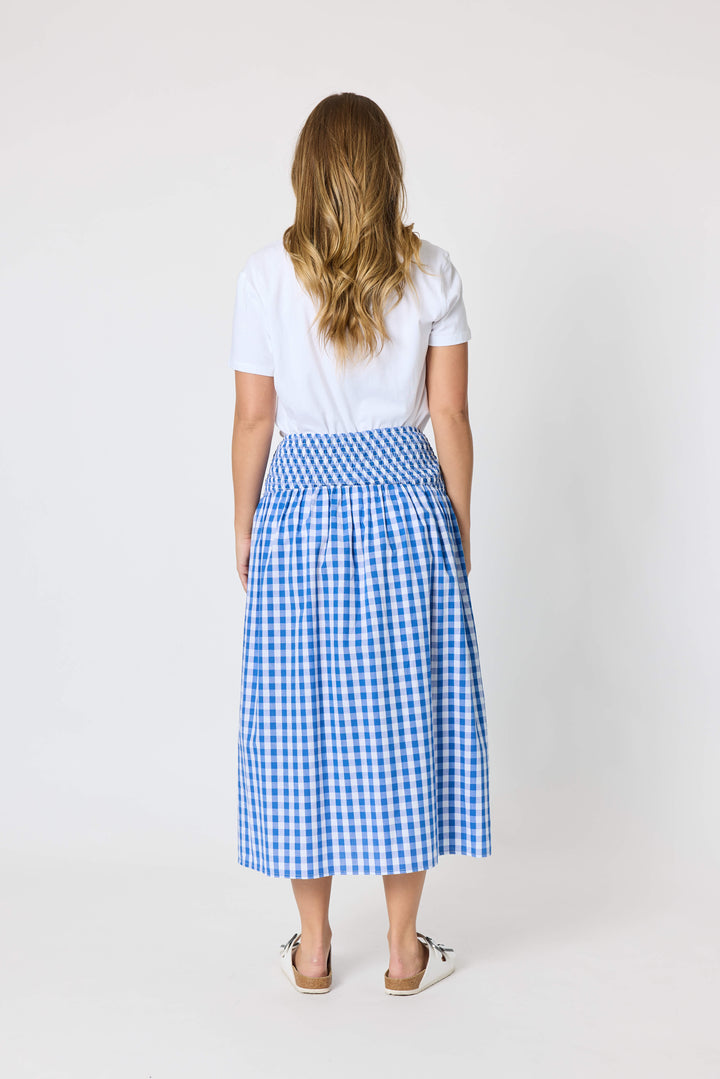 Threadz woman modeling a white graphic t-shirt and a blue and white gingham pleated midi skirt, paired with white slide sandals, against a plain white studio background. Back view
