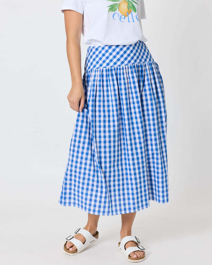 Threadz woman modeling a white graphic t-shirt and a blue and white gingham pleated midi skirt, paired with white slide sandals, against a plain white studio background.