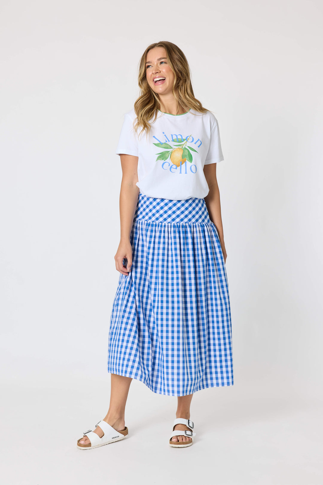 Threadz woman modeling a white graphic t-shirt and a blue and white gingham pleated midi skirt, paired with white slide sandals, against a plain white studio background.