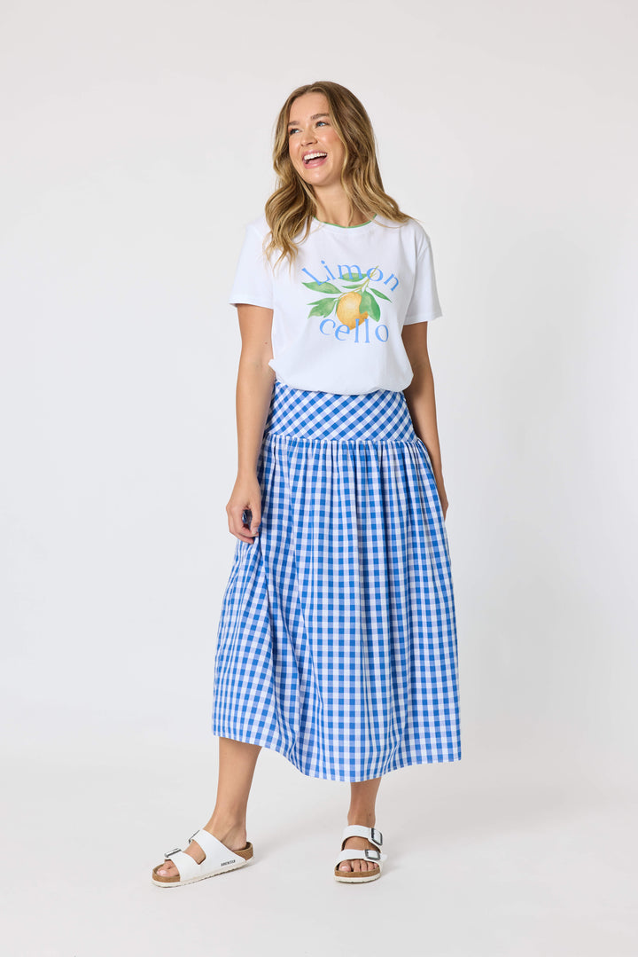 Threadz woman modeling a white graphic t-shirt and a blue and white gingham pleated midi skirt, paired with white slide sandals, against a plain white studio background.