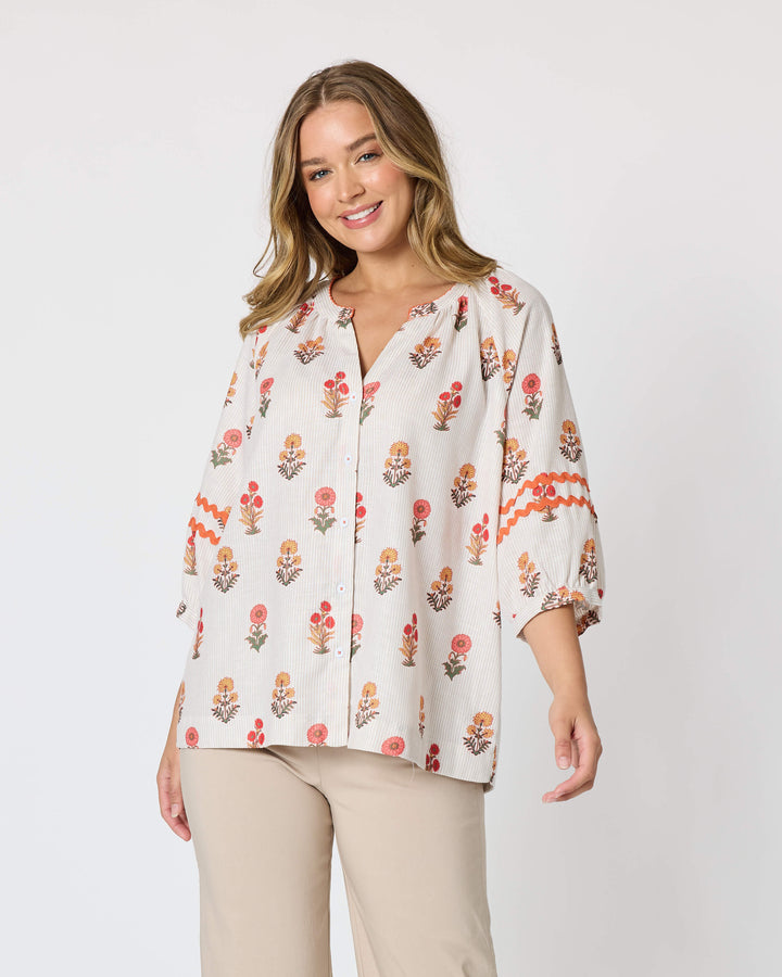Threadz woman modeling a white button-up blouse with a colorful floral and geometric print, paired with coral-colored cropped pants and flat sandals, against a plain white studio background.