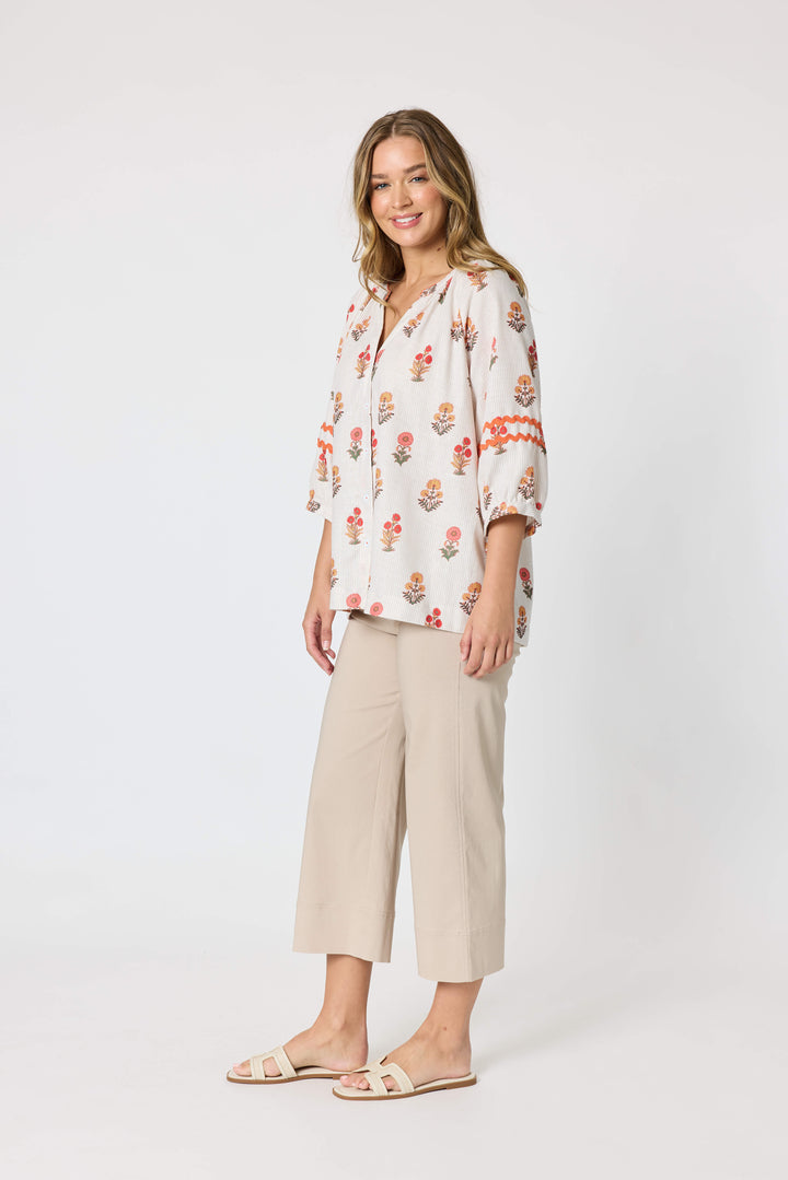 Threadz woman modeling a white button-up blouse with a colorful floral and geometric print, paired with coral-colored cropped pants and flat sandals, against a plain white studio background.