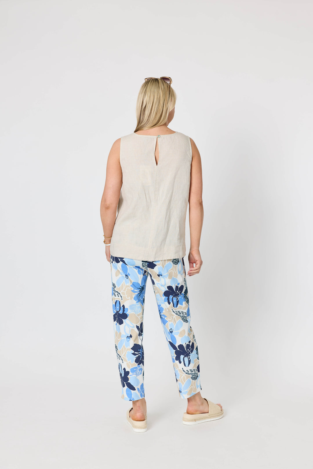 Gordon Smith  woman modeling a sleeveless light beige top and blue floral print pants with cream slide sandals, standing against a plain light gray studio background. Back view