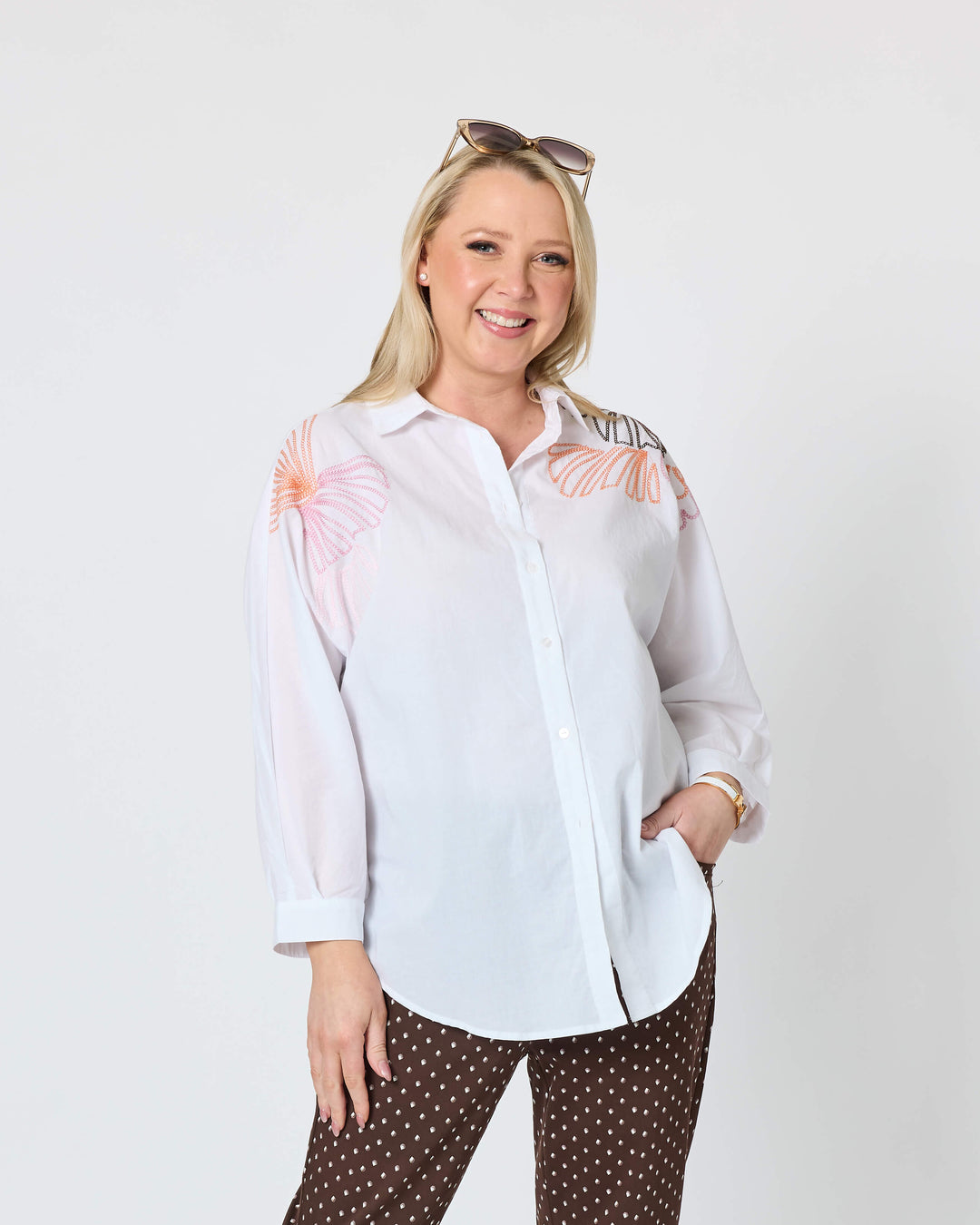 Gordon Smith modeling a white long-sleeve button-up shirt with pink floral embroidery on the shoulders, paired with brown pants featuring a small white geometric pattern, and white slide sandals, against a plain white studio background.