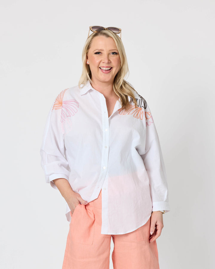 Gordon Smith modeling a white long-sleeve button-up shirt with peach embroidery on the shoulders, paired with peach shorts and white slide sandals, against a plain white studio background.