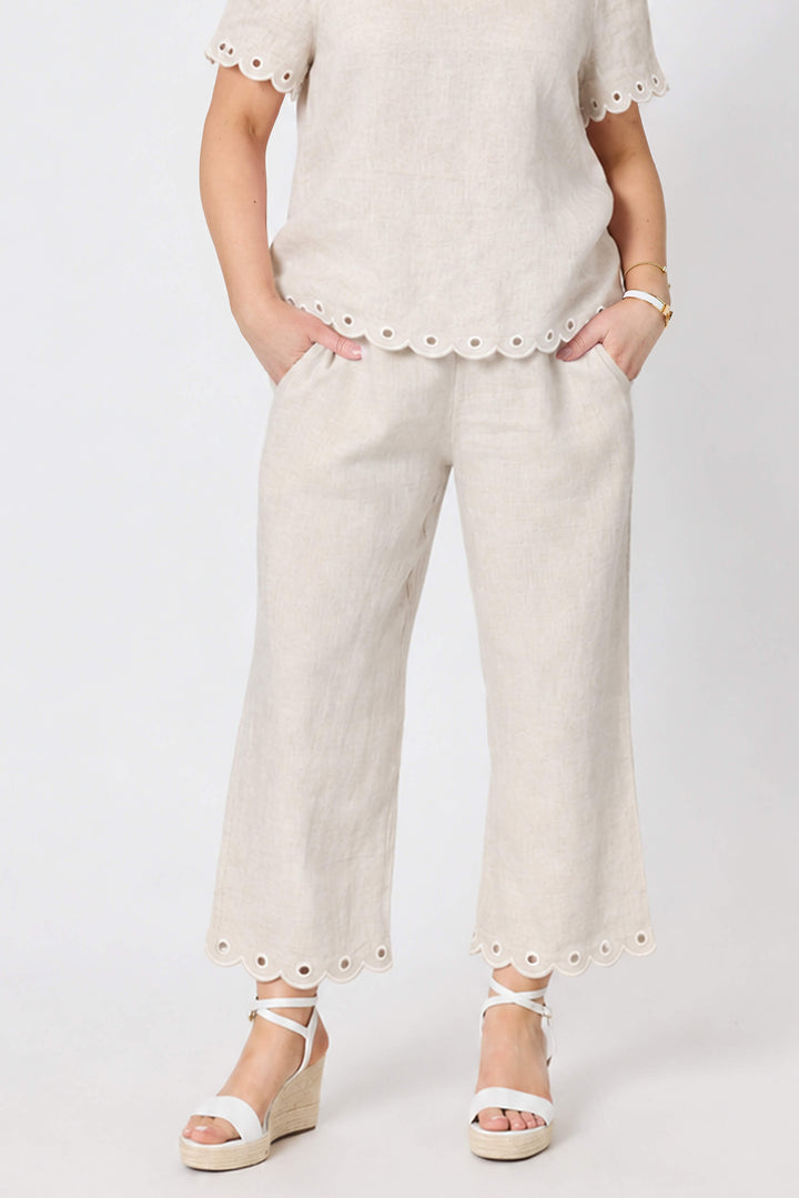 Gordon Smithh  woman modeling a matching beige linen two-piece set consisting of a short-sleeve V-neck top with scalloped edges and matching wide-leg trousers with scalloped hems, styled with white wedge sandals against a light gray studio background.