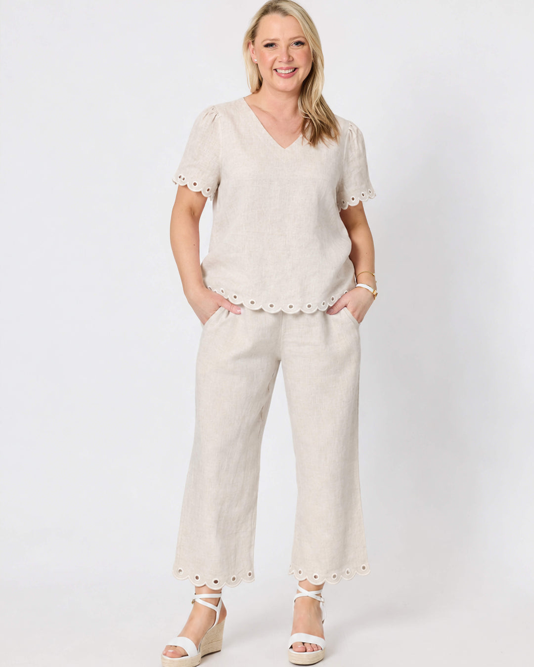 Gordon Smithh  woman modeling a matching beige linen two-piece set consisting of a short-sleeve V-neck top with scalloped edges and matching wide-leg trousers with scalloped hems, styled with white wedge sandals against a light gray studio background.