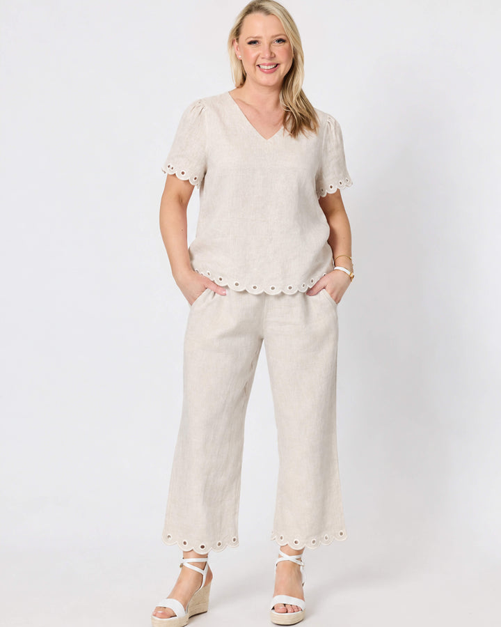 Gordon Smithh  woman modeling a matching beige linen two-piece set consisting of a short-sleeve V-neck top with scalloped edges and matching wide-leg trousers with scalloped hems, styled with white wedge sandals against a light gray studio background.