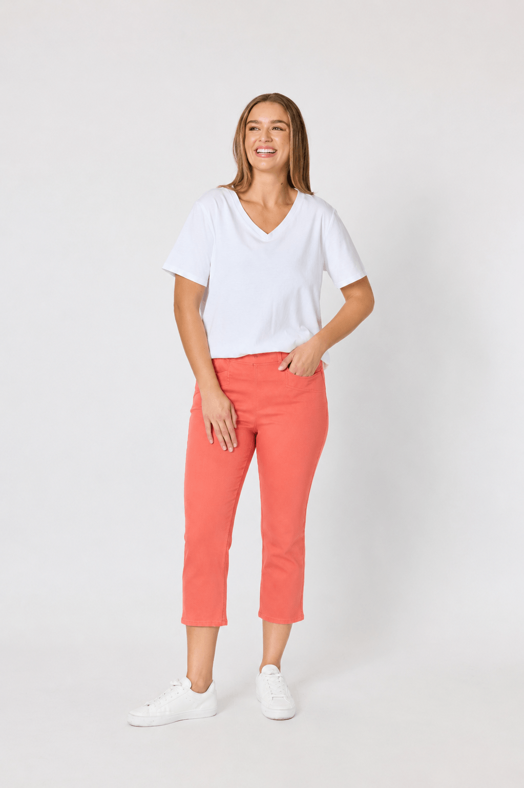 Threadz woman modeling a white V-neck short-sleeve t-shirt and coral orange cropped pants, paired with white sneakers, against a plain light gray studio background.