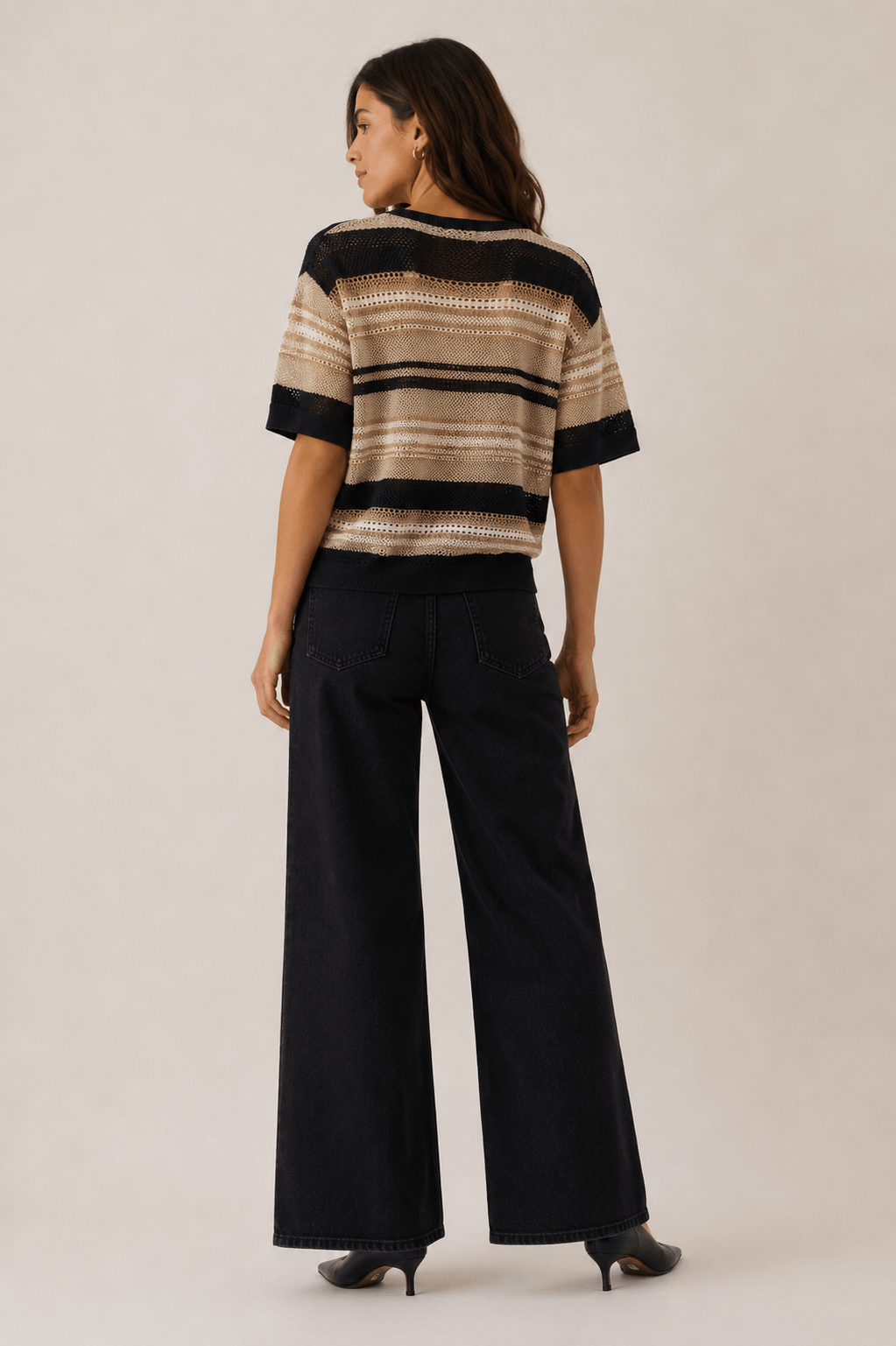 LD&amp;Co modeling wearing a short-sleeve knit sweater with horizontal beige, brown, and black stripes, paired with black jeans, against a plain light beige studio background back view 