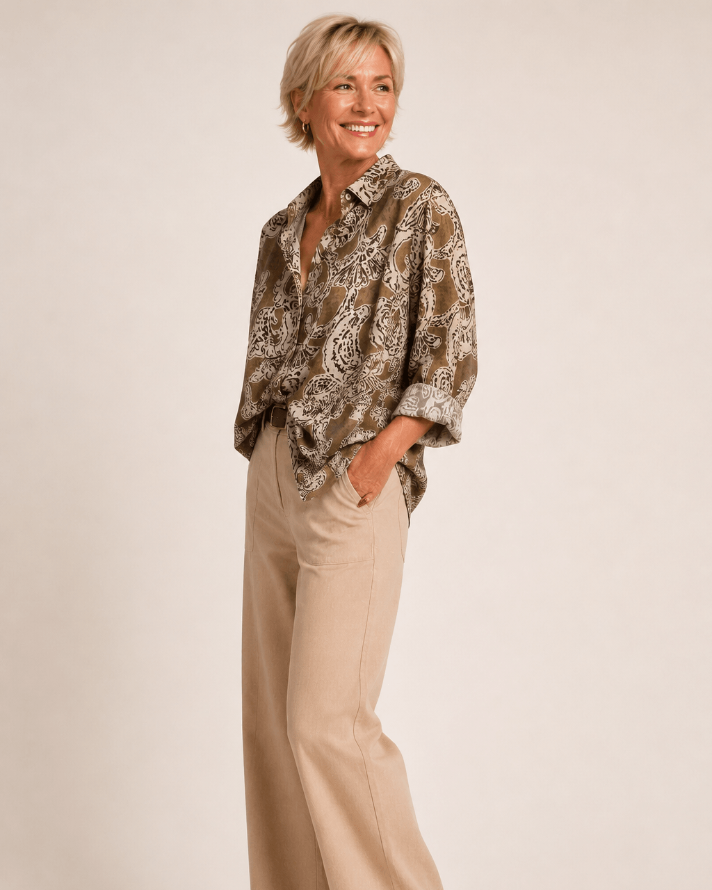 A woman modeling a brown and beige paisley patterned button-down shirt paired with beige wide-leg trousers and brown shoes, standing against a solid light beige studio background.