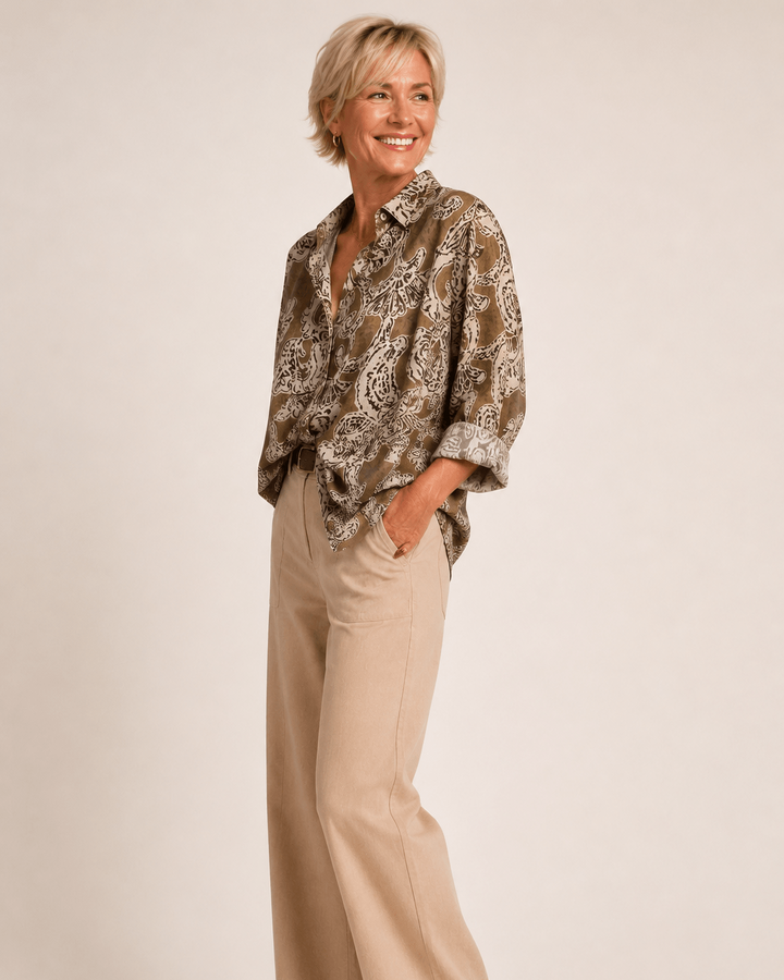 A woman modeling a brown and beige paisley patterned button-down shirt paired with beige wide-leg trousers and brown shoes, standing against a solid light beige studio background.