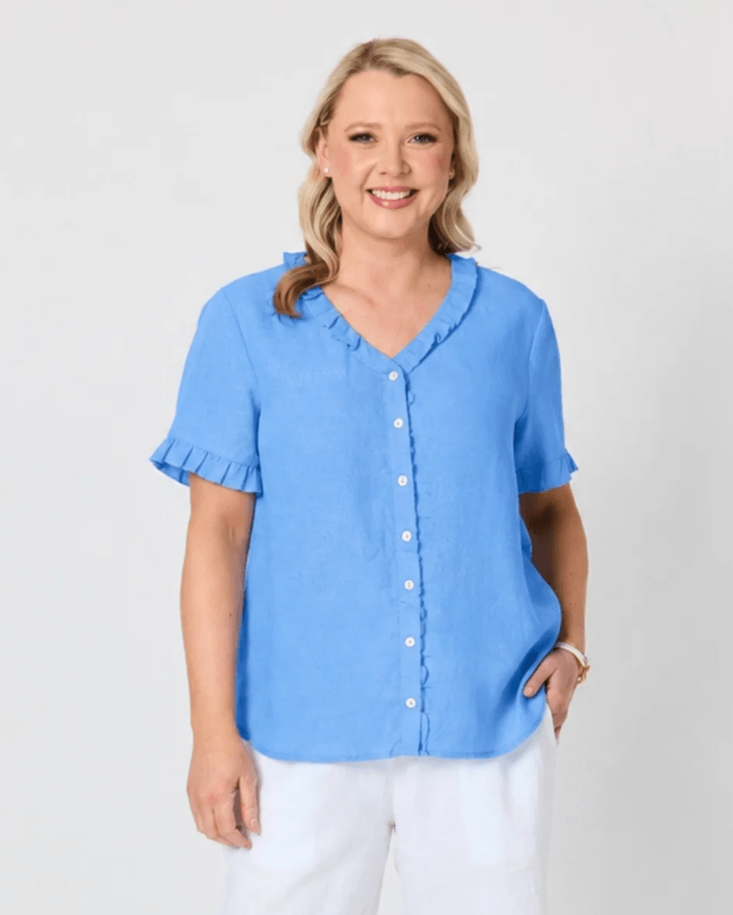 Gordon Smith woman modeling a light blue short-sleeve button-up blouse with ruffled trim at the collar and cuffs, paired with white pants, against a plain light gray studio background.